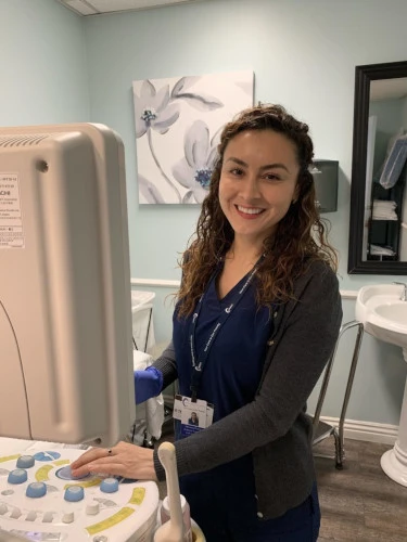 An x-ray technician happily operates the imaging machine, adorned in her professional work uniform. She smiles with confidence, standing beside the advanced x-ray machine, ready to provide quality healthcare services.